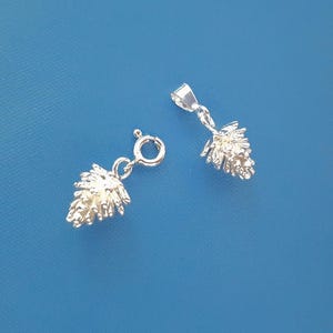 May include: Two silver-toned metal charms shaped like pinecones. One charm features a circular spring ring clasp, while the other charm is attached to a triangular bail, both displaying detailed, textured scales across their surfaces.