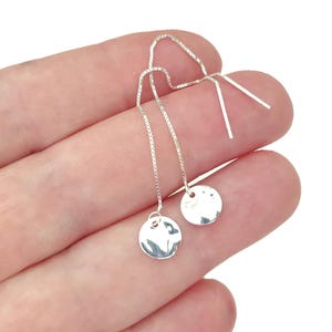 May include: A pair of silver threader earrings featuring delicate box chains that terminate in small, circular, hammered-finish discs. The metallic surface of the silver discs reflects light, creating a bright, polished appearance on the jewellery pieces.