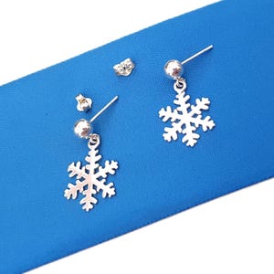 May include: A pair of silver-toned stud earrings featuring a dangling snowflake charm. Each earring consists of a small polished ball post connected to a delicate, intricate snowflake cutout design. Two separate silver-toned earring backs are included.