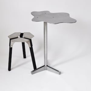 Peut inclure: Une table en métal argenté avec un plateau de forme organique unique et un piètement à trois pieds. Un tabouret en métal noir plus petit avec un piètement à trois pieds est également présent sur l'image.