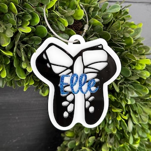 May include: White acrylic ornament shaped like a pair of ballet shoes with a butterfly design. The ornament has the name "Elle" written in blue glitter.