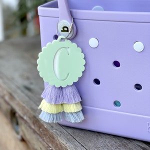 May include: A light purple tote bag with a white circle and a light green circle with the letter "C" cut out. A light purple, yellow, and light blue tassel hangs from the bag.