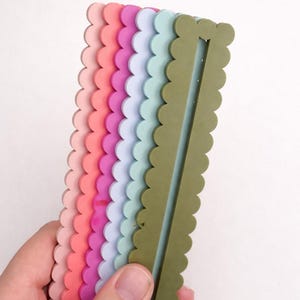 May include: A selection of colourful, scalloped-edge rulers in shades of pink, coral, purple, blue, and green. The rulers are held in a hand against a white background. Each ruler has a central groove.