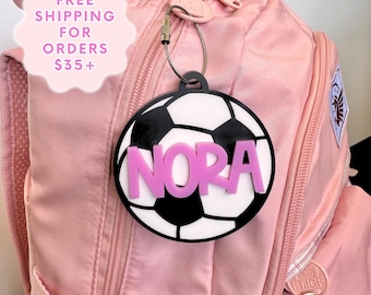May include: A personalized soccer ball luggage tag with the name "NORA" in pink letters. The tag is black and white, attached to a pink backpack. The tag is designed to resemble a soccer ball.