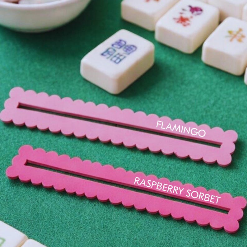 May include: Two pink Mahjong tile holders with scalloped edges, labelled "FLAMINGO" and "RASPBERRY SORBET". The holders are on a green surface, with white Mahjong tiles and a white bowl in the background.