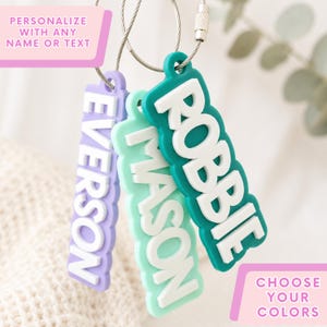 May include: Three rectangular silicone luggage tags hanging from metal wire loops. Each tag features raised white lettering spelling out the names EVERSON, MASON, and ROBBIE in purple, light green, and teal silicone bases respectively.
