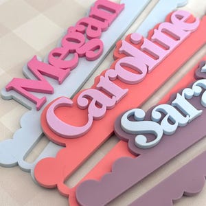 May include: Close-up of personalized name hangers in various pastel colors. The names "Megan", "Caroline", and "Sara" are displayed in layered, 3D lettering. Each hanger has a scalloped edge and a slot for hanging, perfect for a nursery or children's room.