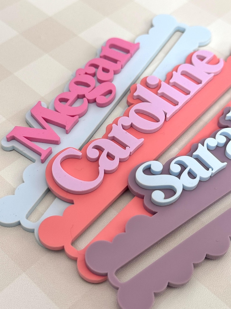 May include: Close-up of personalized name hangers in various pastel colors. The names "Megan", "Caroline", and "Sara" are displayed in layered, 3D lettering. Each hanger has a scalloped edge and a slot for hanging, perfect for a nursery or children's room.