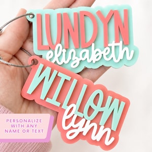 May include: Two custom acrylic luggage tags with a layered design. One tag features the name LUNDYN in coral on a mint base with elizabeth in white script. The other tag features WILLOW in mint on a coral base with lynn in white script.