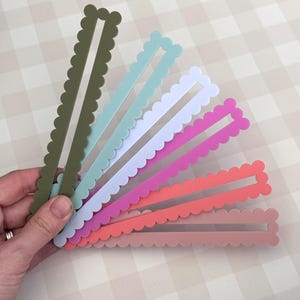 May include: A hand holds a fan of colourful, scalloped-edge, rectangular craft pieces. The colours include olive green, light blue, white, pink, coral, and a muted rose. Each piece has a rectangular cutout in the centre.