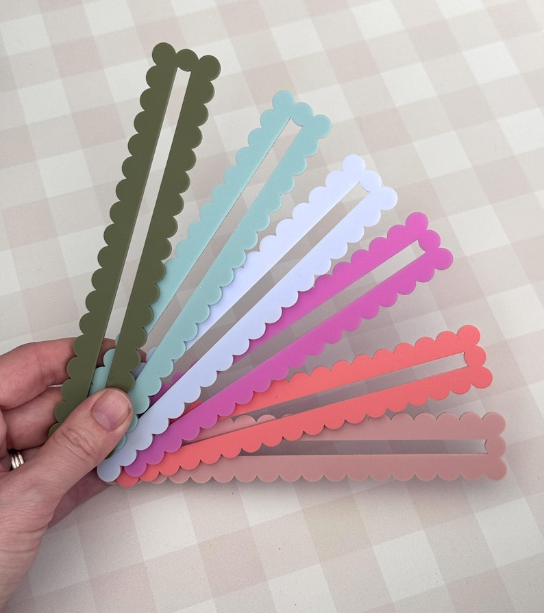 May include: A hand holds a fan of colourful, scalloped-edge, rectangular craft pieces. The colours include olive green, light blue, white, pink, coral, and a muted rose. Each piece has a rectangular cutout in the centre.