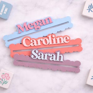 May include: Three personalized name holders in pastel colors: light blue, coral, and lavender, each with a name in a contrasting color. Mahjong tiles with various designs surround the name holders.