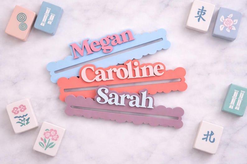 May include: Three personalized name holders in pastel colors: light blue, coral, and lavender, each with a name in a contrasting color. Mahjong tiles with various designs surround the name holders.
