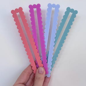 May include: Four colourful hair clips with a scalloped edge, in coral, purple, light blue, and teal. The clips are held in a hand against a white background. Each clip has a rectangular centre.