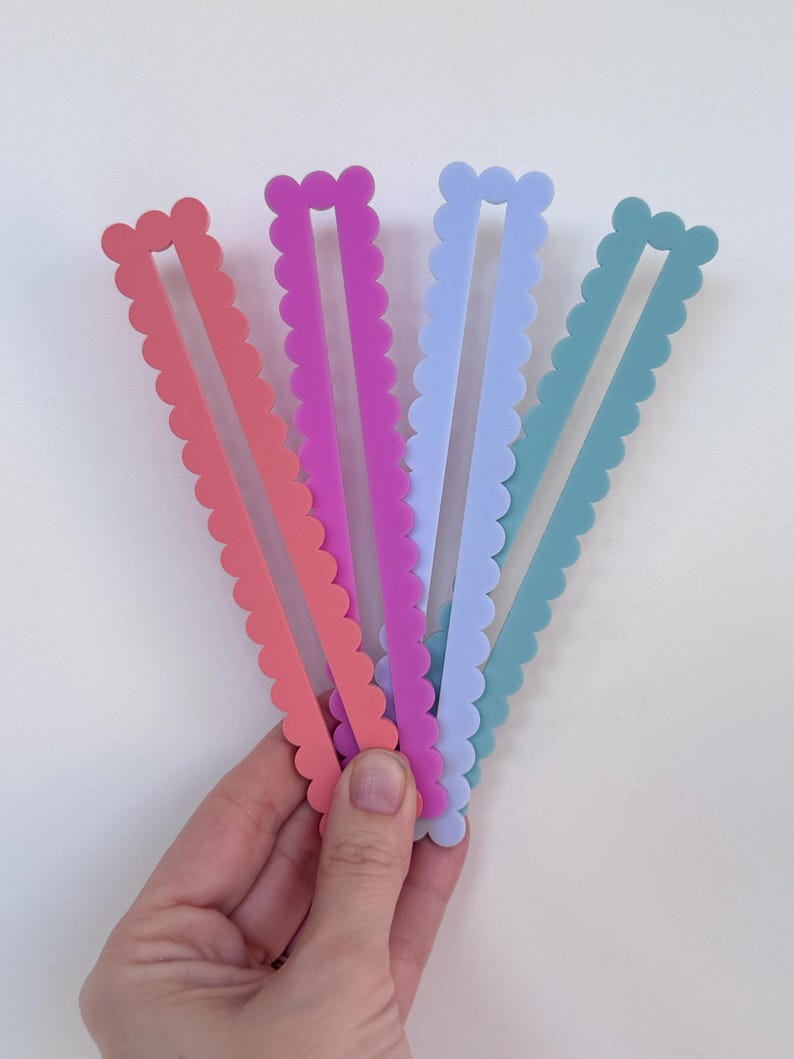 May include: Four colourful hair clips with a scalloped edge, in coral, purple, light blue, and teal. The clips are held in a hand against a white background. Each clip has a rectangular centre.