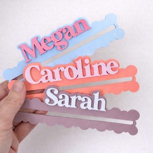 May include: Three personalized name tags in pastel colors: blue with "Megan" in pink, coral with "Caroline" in pink, and mauve with "Sarah" in white. Each tag has a cloud-shaped border.