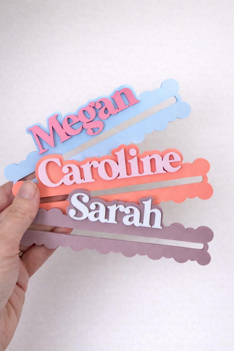 May include: Three personalized name tags in pastel colors: blue with "Megan" in pink, coral with "Caroline" in pink, and mauve with "Sarah" in white. Each tag has a cloud-shaped border.