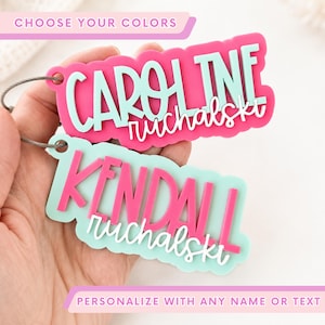 May include: Two custom acrylic name tags with a layered design. The top tag features the name CAROLINE in mint green on a hot pink background with the white script text ruchalski. The bottom tag features KENDALL in hot pink on a mint green background with the white script text ruchalski. Both tags include a metal wire loop.