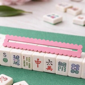 May include: A Mahjong set with white tiles, each displaying Chinese characters and symbols. A pink, scalloped-edge wooden piece sits atop the tiles. The background features a green surface, pink flowers, and a pair of glasses.