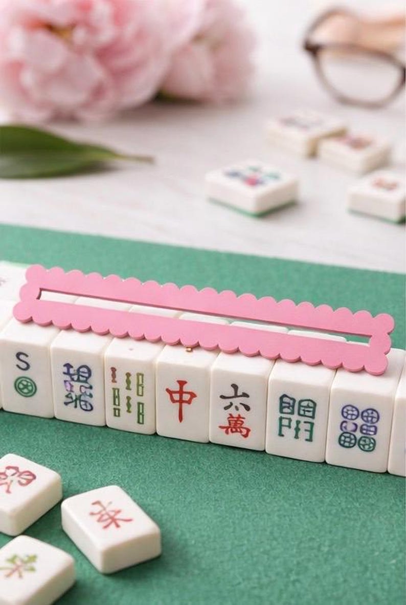 May include: A Mahjong set with white tiles, each displaying Chinese characters and symbols. A pink, scalloped-edge wooden piece sits atop the tiles. The background features a green surface, pink flowers, and a pair of glasses.