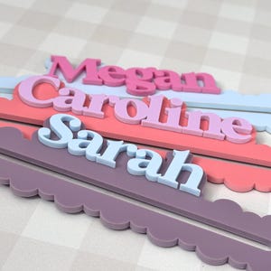 May include: Three personalized name signs in a layered design. The names "Megan", "Caroline", and "Sarah" are displayed in different colors and fonts. Each name is set on a scalloped edge base in coordinating colors.