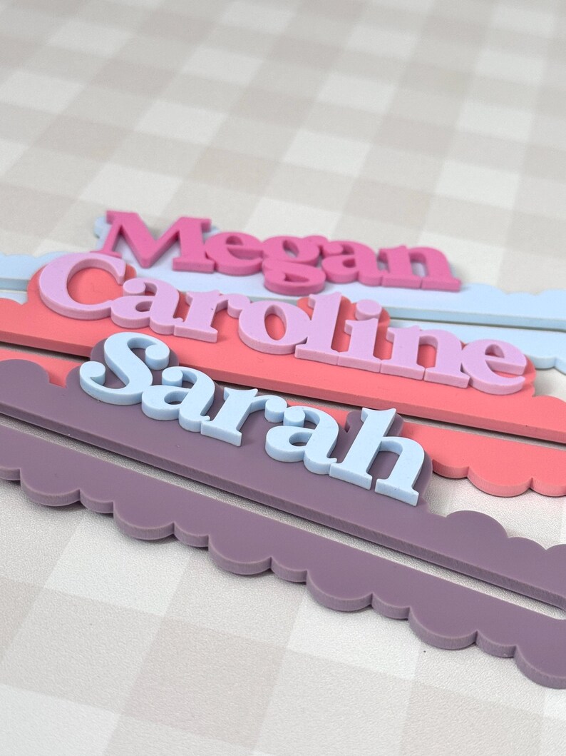 May include: Three personalized name signs in a layered design. The names "Megan", "Caroline", and "Sarah" are displayed in different colors and fonts. Each name is set on a scalloped edge base in coordinating colors.