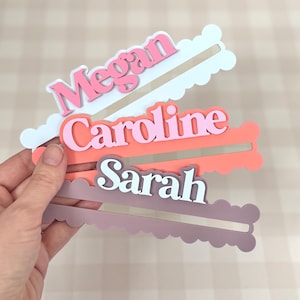 May include: Three personalized name tags in pink, coral, and mauve, each with a unique name: "Megan", "Caroline", and "Sarah". The names are in a contrasting color from the background. The tags have a cloud-like shape with a slot for insertion.