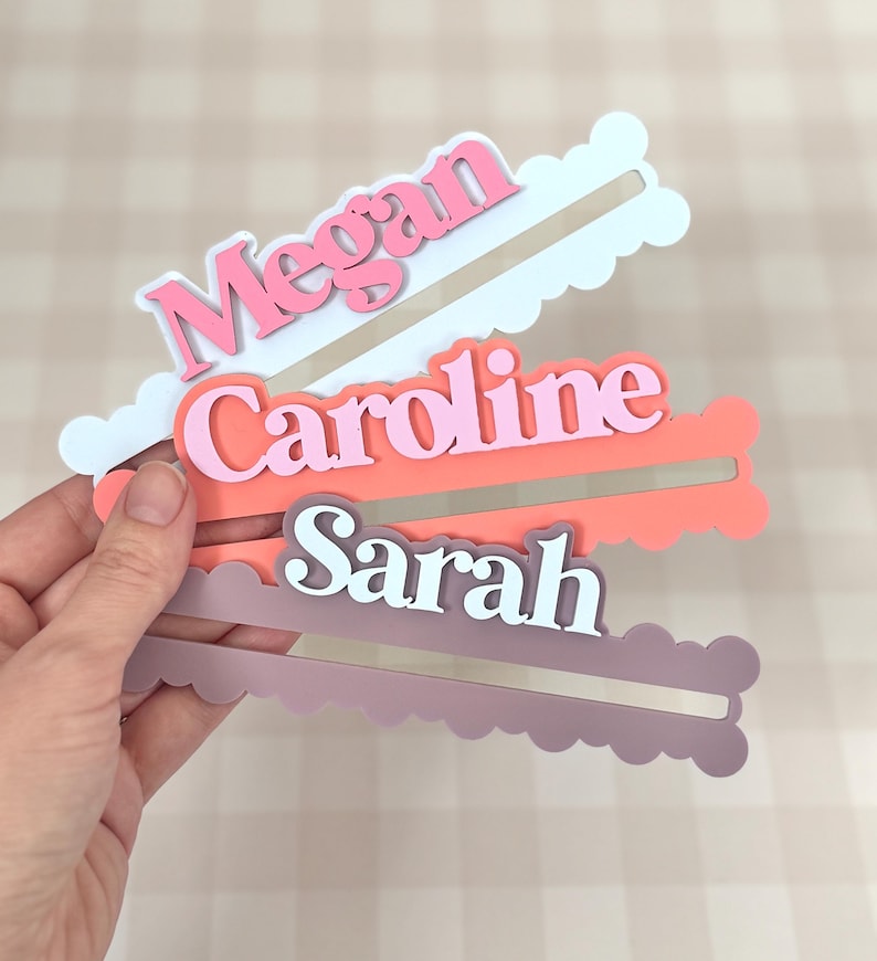 May include: Three personalized name tags in pink, coral, and mauve, each with a unique name: "Megan", "Caroline", and "Sarah". The names are in a contrasting color from the background. The tags have a cloud-like shape with a slot for insertion.