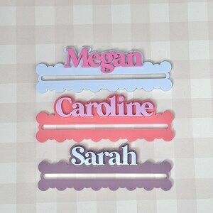 May include: Three personalized name cutouts: "Megan" in pink on a light blue base, "Caroline" in pink on a coral base, and "Sarah" in white on a purple base. Each name is in a script font and set on a scalloped edge base.