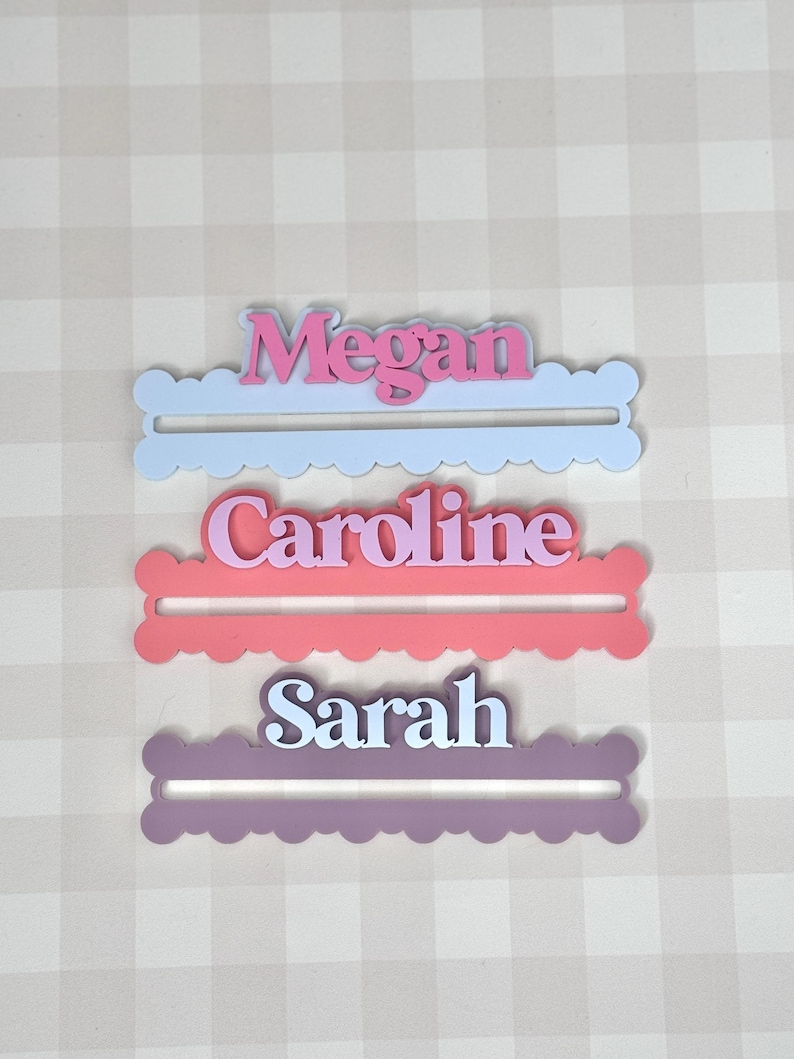 May include: Three personalized name cutouts: "Megan" in pink on a light blue base, "Caroline" in pink on a coral base, and "Sarah" in white on a purple base. Each name is in a script font and set on a scalloped edge base.
