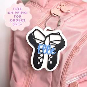 May include: A white and black luggage tag with a ballet shoe design and the name "Elle" in blue script. The tag is attached to a pink backpack with a silver wire loop.