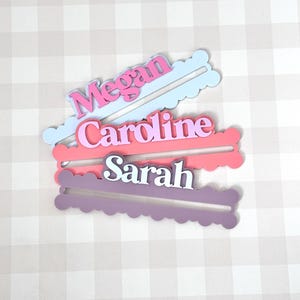 May include: Three layered name tags in pink, purple, and blue. Each tag has a name: Megan, Caroline, and Sarah. The names are in white, with a colored outline. The tags have a scalloped edge and a slot for placement.