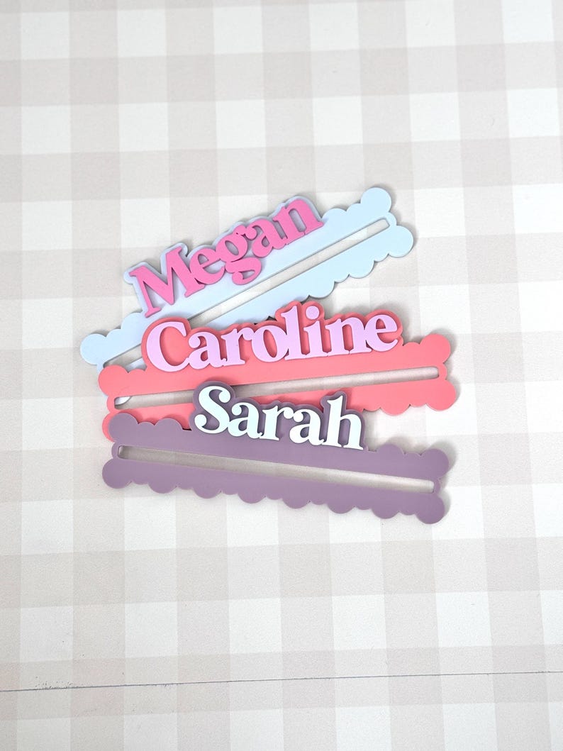 May include: Three layered name tags in pink, purple, and blue. Each tag has a name: Megan, Caroline, and Sarah. The names are in white, with a colored outline. The tags have a scalloped edge and a slot for placement.