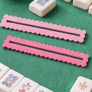 May include: A close-up of a Mahjong game in progress, set on a green surface. Two pink, scalloped tile racks are in the foreground. White Mahjong tiles with colourful symbols and characters are scattered around. A small white bowl with white game pieces is in the background.