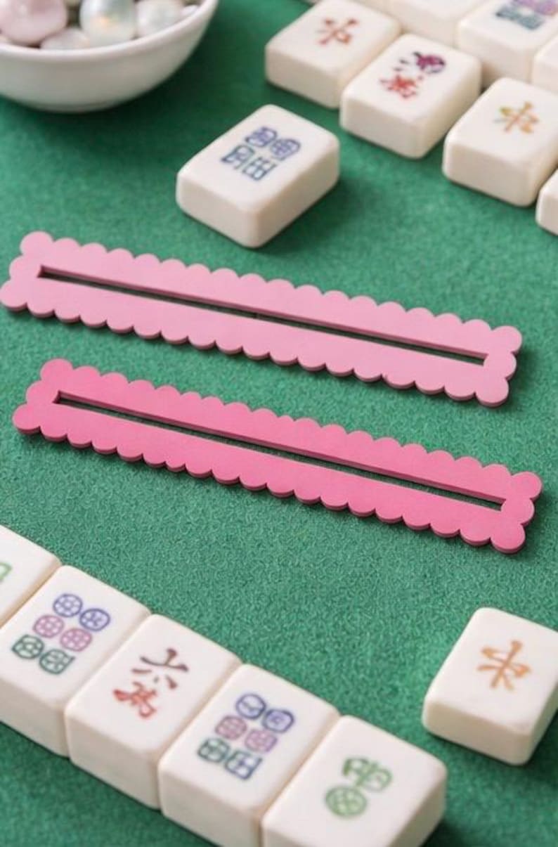May include: A close-up of a Mahjong game in progress, set on a green surface. Two pink, scalloped tile racks are in the foreground. White Mahjong tiles with colourful symbols and characters are scattered around. A small white bowl with white game pieces is in the background.