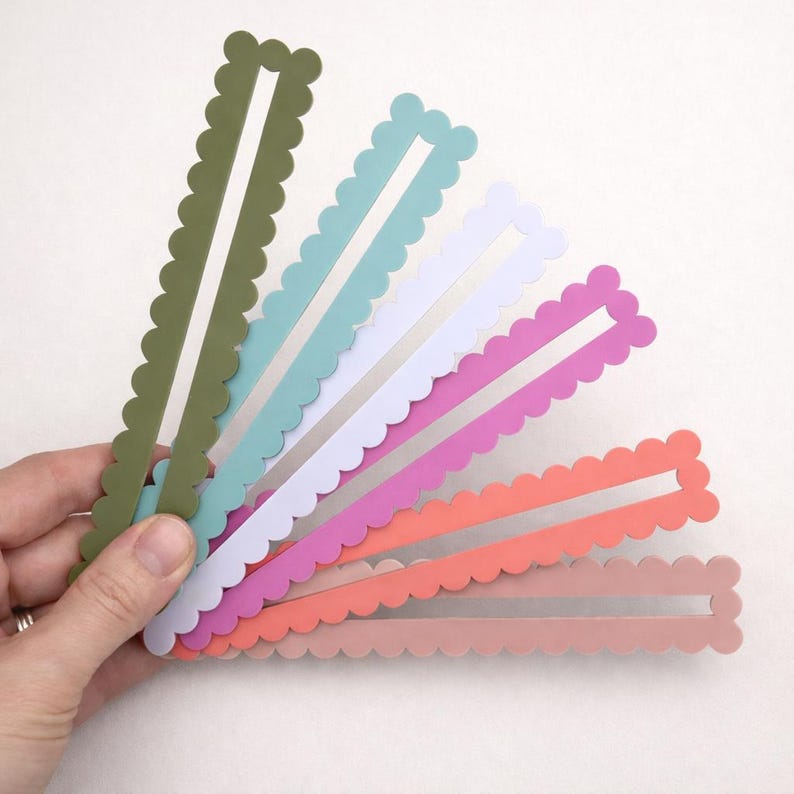 May include: A hand holds a fan of colourful paper strips. Each strip has a scalloped edge and a rectangular cutout. Colours include olive green, teal, white, pink, coral, and peach.