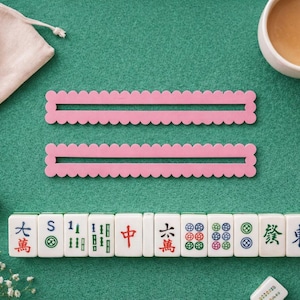 May include: Two pink, scalloped-edge Mahjong tile racks and a row of Mahjong tiles are arranged on a green felt surface. A small white pouch and a cup of tea are also visible. The tiles have Chinese characters and symbols.