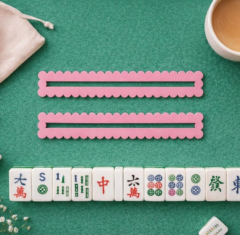 May include: Two pink, scalloped-edge Mahjong tile racks and a row of Mahjong tiles are arranged on a green felt surface. A small white pouch and a cup of tea are also visible. The tiles have Chinese characters and symbols.
