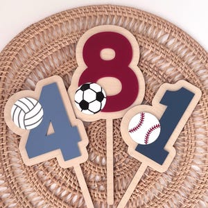 May include: Wooden number toppers for a birthday cake. The numbers are 4, 8, and 1, with sports ball accents. The number 4 is blue with a volleyball. The number 8 is maroon with a soccer ball. The number 1 is blue with a baseball.
