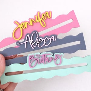 May include: Three personalized name place cards in pink, gray, and mint green. Each card has a wavy edge and a slot to hold a name. The names "Jennifer", "Alissa", and "Brittany" are displayed in a cursive font.