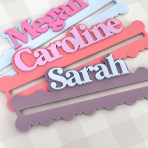 May include: Three personalized name tags in pastel colors: pink, coral, and lavender. Each tag features a name in a contrasting color, with the names "Megan", "Caroline", and "Sarah" displayed in a playful font. The tags have a scalloped edge.