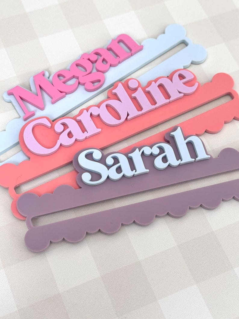 May include: Three personalized name tags in pastel colors: pink, coral, and lavender. Each tag features a name in a contrasting color, with the names "Megan", "Caroline", and "Sarah" displayed in a playful font. The tags have a scalloped edge.