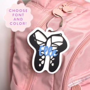 May include: A black and white ballet slipper shaped bag tag with the name Elle in light blue script lettering attached to a pink backpack with a metal wire loop. The image includes the text CHOOSE FONT AND COLOR in a pink scalloped bubble.