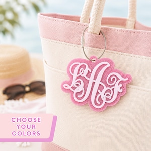 May include: A pink glittery acrylic monogram tag featuring the letters LAF in a cursive script, attached to a cream and pink canvas tote bag with a silver metal cable ring. The tag has a scalloped edge and a smooth light pink letter overlay.