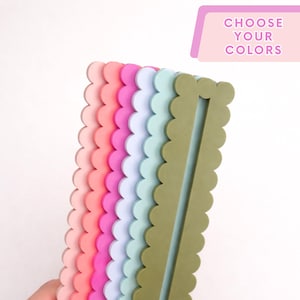 May include: A stack of six rectangular silicone bookmarks with scalloped edges in olive green, light blue, lavender, magenta, coral, and light pink. The top bookmark features a vertical cutout slot. Text at the top reads CHOOSE YOUR COLORS.