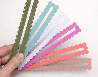 May include: A hand holds a fan of colorful paper strips. Each strip has a scalloped edge and a rectangular cutout. Colors include olive green, teal, white, pink, coral, and peach.