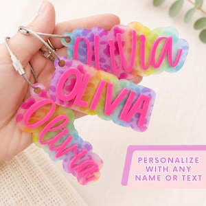 May include: Three custom acrylic keychains featuring a rainbow-colored leopard print background and the name Olivia in raised, bright pink script lettering. Each keychain is attached to a flexible silver-toned metal cable loop for secure attachment.