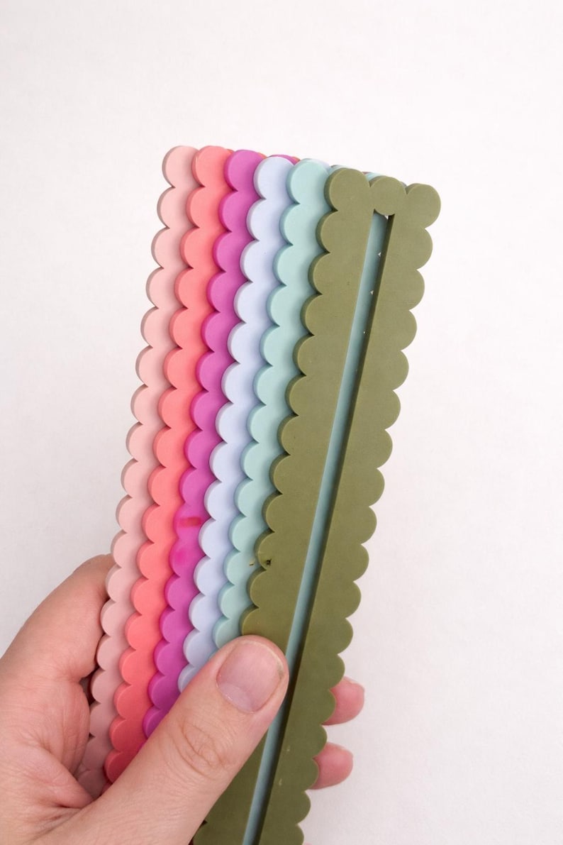 May include: A selection of colourful, scalloped-edge rulers in shades of pink, coral, purple, blue, and green. The rulers are held in a hand against a white background. Each ruler is approximately 15 cm in length.