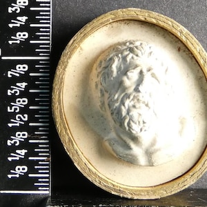 Pode incluir: Escultura em relevo ovalada em estilo antigo, apresentando um busto masculino clássico. O busto é branco, sobre um fundo creme, e emoldurado por uma borda dourada texturizada. A peça mede aproximadamente 2,5 cm de diâmetro.