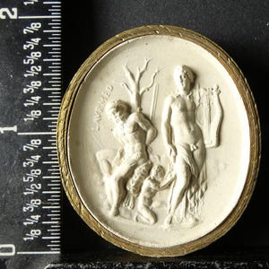 Puede incluir: Escultura en relieve ovalada, blanquecina, con marco de metal dorado. El relieve representa figuras clásicas, incluido un hombre con una lira. La palabra "LAVRMED" está inscrita. La pieza mide aproximadamente 5 cm de diámetro.